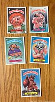 Front view of five Garbage Pail Kids stickers showing characters Mogged Marcus, Boogers Sean, Broad Maud, Bony Tony, and Alice Island with colorful grotesque artwork.