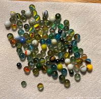 Photo showing approximately 87 assorted glass marbles of various colors and sizes on a beige carpeted surface.