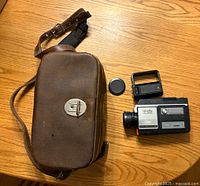 Photo of Minolta XL-250 camera with lens cover, detachable viewfinder, and brown leather carrying case with shoulder strap displayed on wooden surface.