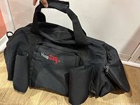 Front view of the black HoopSaq basketball gym bag showing two cylindrical holders, front zipped pouch with HoopSaq logo, and carry handles.