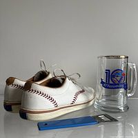 Rear-side angle of vintage Keds baseball shoes with visible red baseball stitch design on white leather, 10th anniversary Blue Jays glass mug, and blue-handled Old Vienna bottle opener placed in front.