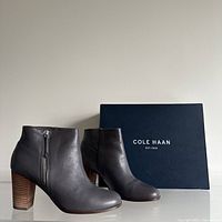 Photo of the pair of Cole Haan leather ankle boots shown with their original navy blue box. Boots are dark brown with side zippers and wooden block heels.