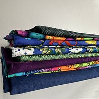 Stack of multiple types of fabric with different colorful prints and solid colors including polka dots and floral patterns, folded and stacked against a white wall.