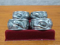 Four pewter napkin rings with high-relief Haida motifs displayed on a red velvet presentation box, showing front and side views of the detailed whale and calf designs.