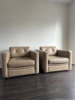 Front view showing the sable/taupe leather club chair with a tufted backrest and cushioned seat, placed next to a matching chair.