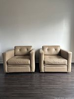 Front view of two identical beige leather club chairs with button tufted backs and wide square arms on wooden floor.