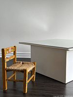 Side angled view of the hand painted Mexican child's chair showing painted floral designs, woven seat, and proximity to a white table for scale.