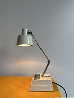 Small cream and chrome mid-century modern desk lamp on yellow surface against white wall. Lamp head is angled downward and lit.