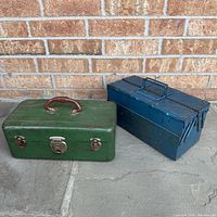 Pair of two metal storage boxes placed on concrete ground against a brick wall background. The green box is on the left and the blue box on the right.