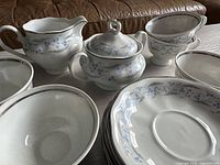Close-up view of the creamer, lidded sugar bowl, teacups and saucers arranged on a table with a leather couch in background