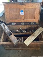 Open antique wooden chest showing two removable wooden trays holding various carpentry tools including wooden planes, a metal hand plane, and a spirit level.
