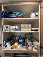 Wooden shelving unit with assorted household items including reusable bags on top shelf, pill boxes and lightbulbs on second shelf, assorted household supplies on bottom shelf including feather duster.