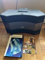 Bose Acoustic Wave Music System with accompanying remote controls and owner's manual laid out on floor showing full system and documents.