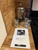 Photo of the Maxim French Press, electric warming base, two clear glass cups, and instruction manual on a wooden countertop.