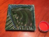 Top view of green ceramic Royal Haegar horse head ashtray and small round orange and chrome ashtray on wooden surface