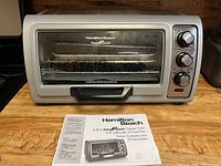 Front view of the silver and black Hamilton Beach Easy Reach 6 slice toaster oven with manual placed in front.