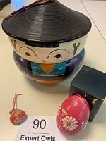 Front view of stacked Kokeshi bento bowls with conical hat lid, dark pink hand-painted eggshell ornament in original box, and kimono fabric charm with red string and bell