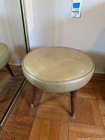 Front angle of circular padded foot stool with beige vinyl upholstery and wooden legs with brass feet on parquet floor, next to mirrored closet door.