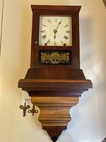 Front view of wooden pillar wall clock showing painted dial with Roman numerals, decorative glass panel, and two brass keys attached.