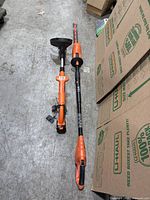 Full view showing two cordless Black and Decker orange trimmers placed on ground by boxes.