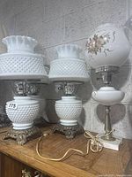 Four vintage electric lamps displayed on a wooden surface against a concrete block wall. Three are white milk glass hobnail style lamps with ruffled tops and ornate brass bases. Fourth lamp is white milk glass with floral print globe shade and marble base.