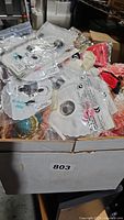 Box open showing various doll items including packaged doll eyes, fabric scraps, and small clothing pieces alongside boxed and loose accessories