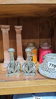 Pair of metal bird motif candlesticks, pink column candlesticks, three scented candles in glass jars, and mosaic glass candle holder on wooden shelf.