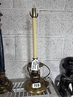 Full view of the Chapman brass candlestick lamp with a fitted candle-shaped light bulb holder, standing about 20 inches tall against a gray cinderblock wall.