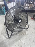 Full view of black metal floor fan on concrete floor. Shows the metal cage, three blades, and supporting frame.