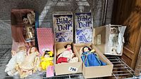 Full lot of vintage dolls arranged on a wire shelf; includes two boxed Bible Dolls and several other assorted vintage dolls in dresses and other vintage clothing.