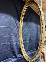 Full view of the oval gold gilt mirror showing the shape and detailed frame design against a black fabric background.