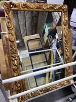 Full view of the large rectangular mirror with ornate gold gilt frame leaning on storage rack showing carved floral and scroll design.