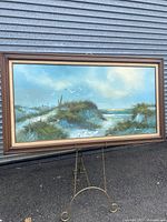 Full view of framed seascape painting displayed on metal stand outdoors, showing coastal scene with ocean, dunes, grasses, and birds.