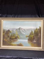 Full view of framed oil on canvas landscape painting showing mountain, river, trees, and cliffs in a naturalistic style with muted colors.