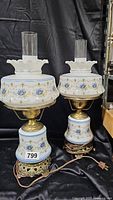 Pair of Abigail Adams hurricane lamps with blue poppy floral design on white glass, brass bases, glass chimneys, and ornate metal feet shown against black backdrop.