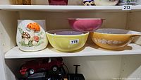 Four Pyrex mixing bowls in different colors and a mushroom-themed ceramic canister on a shelf. The bowls include pink, purple, yellow, and orange with white floral designs. The ceramic canister has a raised design of mushrooms painted in orange, brown, and red hues with green foliage.
