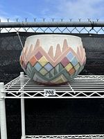 Side view of the multi-colored pastel Southwest Aztec ceramic planter on a white wire shelf with chain link fence background.