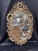 Front view of ornate oval mirror with intricate gold-tone frame showing scroll and floral patterns, displayed against black cloth backdrop.