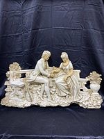 Full view of the colonial couple wall hanging with the man proposing to the woman on a bench surrounded by sculpted foliage and a decorative urn.