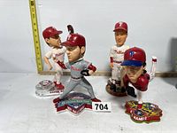 Overall group photo of four Phillies bobbleheads including Aaron Rowand, Roy Halladay, Richie Ashburn, and Cole Hamels on table with measuring tape in background.