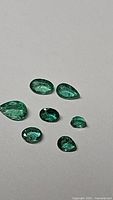 Seven loose faceted gemstones of Colombian emeralds in various cuts, showing their rich green color and clarity.