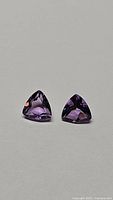 Pair of natural purple amethyst gemstones, trillion cut, placed side by side on a plain background showing shape and color.