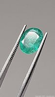 Close-up view of the oval cut emerald held with tweezers against a neutral background to highlight the gemstone's color and cut.