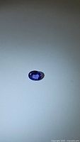 Oval cut deep purple-blue tanzanite gemstone under light, showing color and facets clearly against plain background.