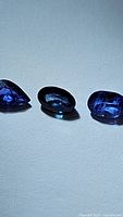 Three blue sapphires showing pear, oval, and cushion cuts and rich blue color with internal characteristics visible under lighting.