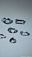 Six different shaped aquamarine gemstones arranged on a flat surface with lighting highlighting the facets and clarity.