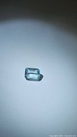 Photograph of a natural sky blue aquamarine gemstone, cut in emerald shape with visible facets and high clarity, placed on white background under focused light.