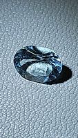Close-up photo of a natural aquamarine oval cut gemstone showing its light blue color, facets and clarity on a textured surface.