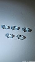 Photo showing five oval cut aquamarine gemstones arranged in two rows on a grey surface, displaying their clarity and light blue color under lighting.