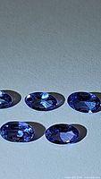 Five natural calibrated oval cut tanzanite gemstones displayed on a plain surface with visible facets and purple-blue color.